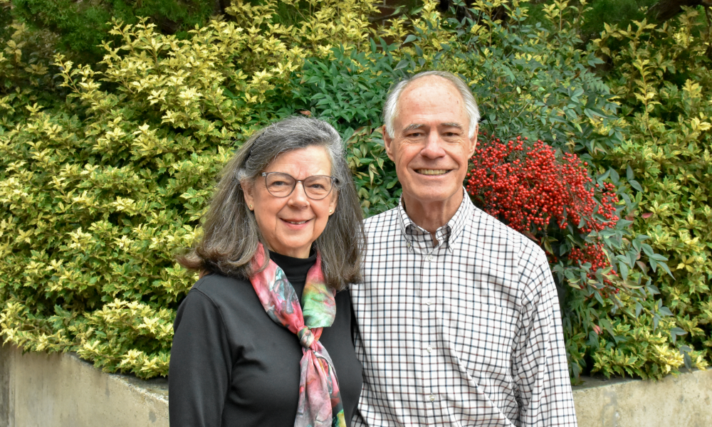A man and woman smile in front of a green and red hedge